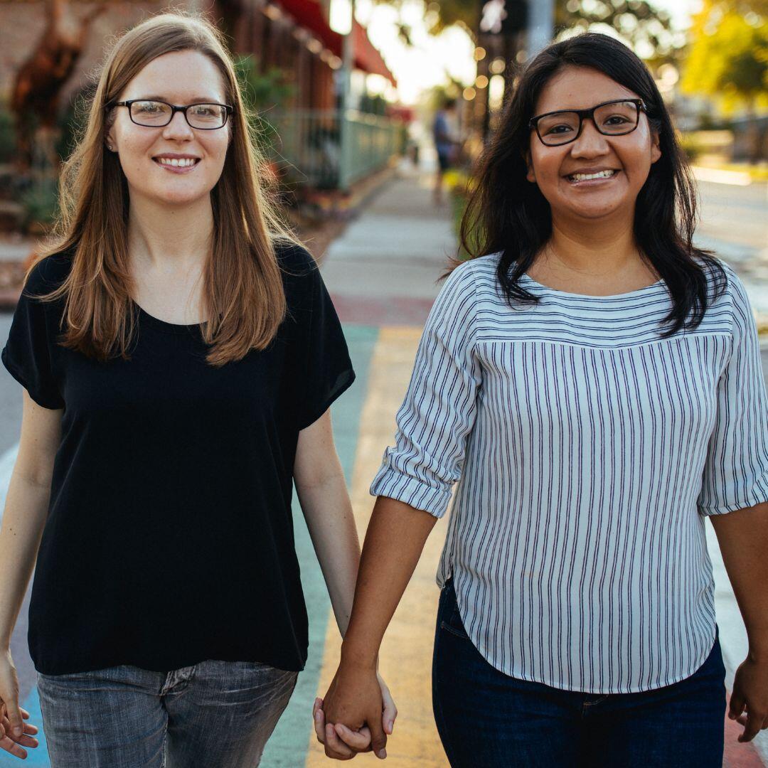 LGBT couple looking to adopt in Houston, TX (white female and latino female, both with long hair and glasses) holding hands